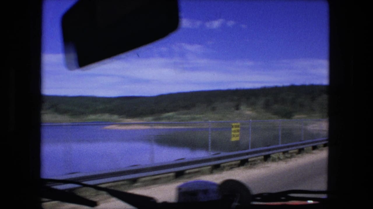 View through a vehicle windshield toward a fenced lakeside road and low hills.