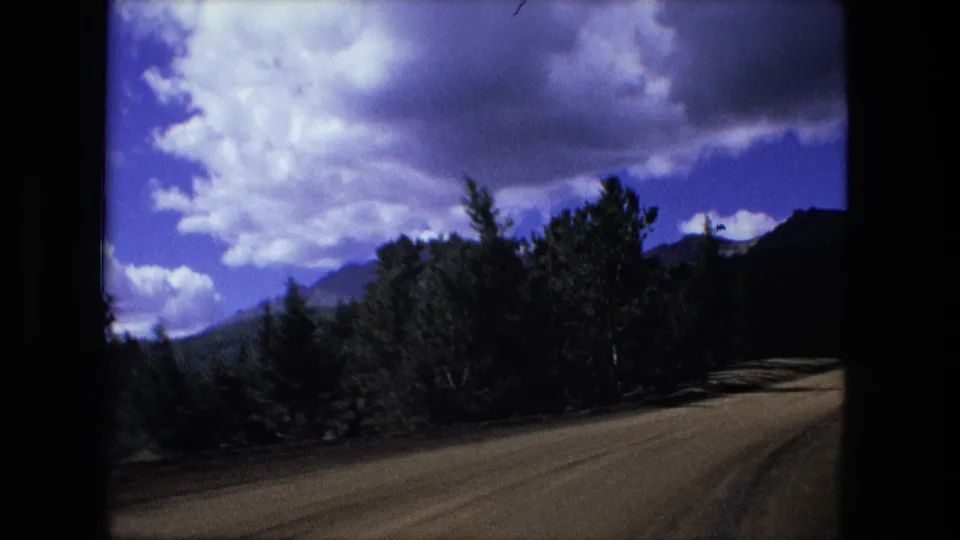 1976 8mm travel footage shows a dusty rural road lined with trees, with dark mountain forms and dramatic clouds beyond.