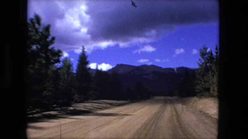 1976 8mm home-movie frame of a dirt road running through pine trees toward distant mountains beneath a vivid blue, clouded sky.