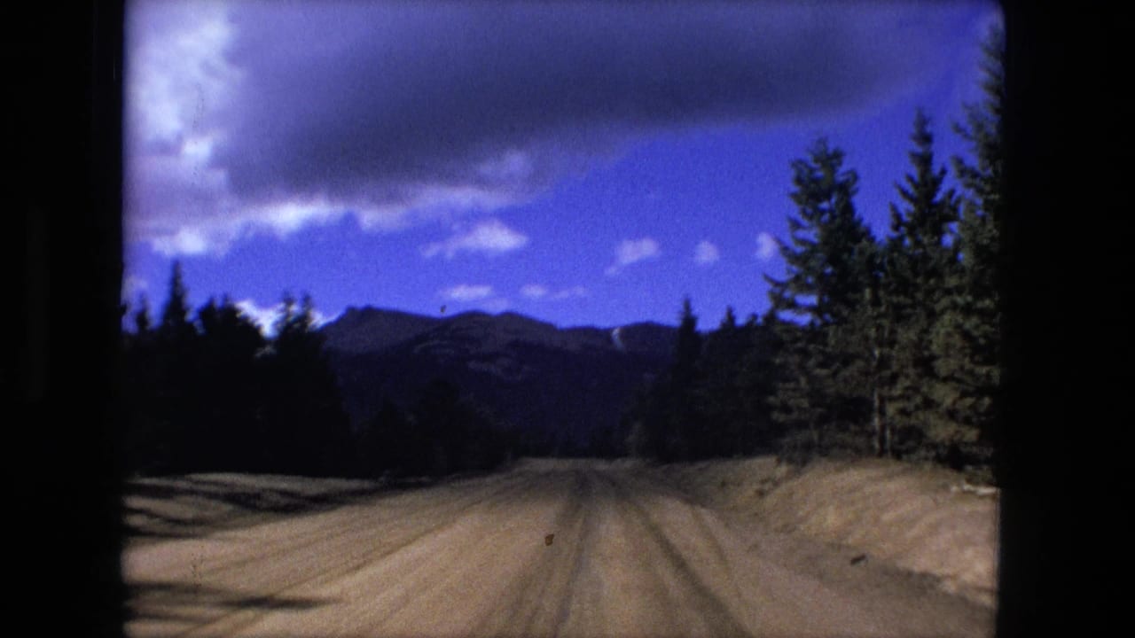Unpaved mountain road lined with evergreen trees under a blue cloudy sky.