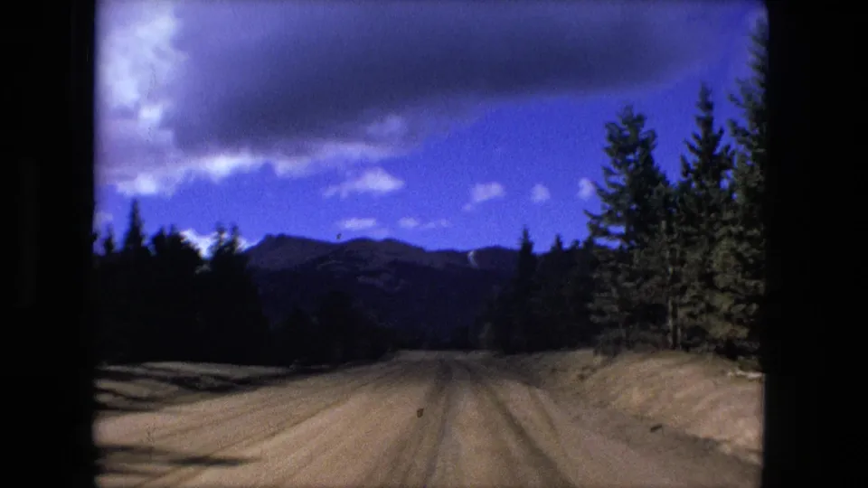 1976 8mm frame of a forest road with evergreen trees, distant mountain ridges, and a blue sky with clouds.