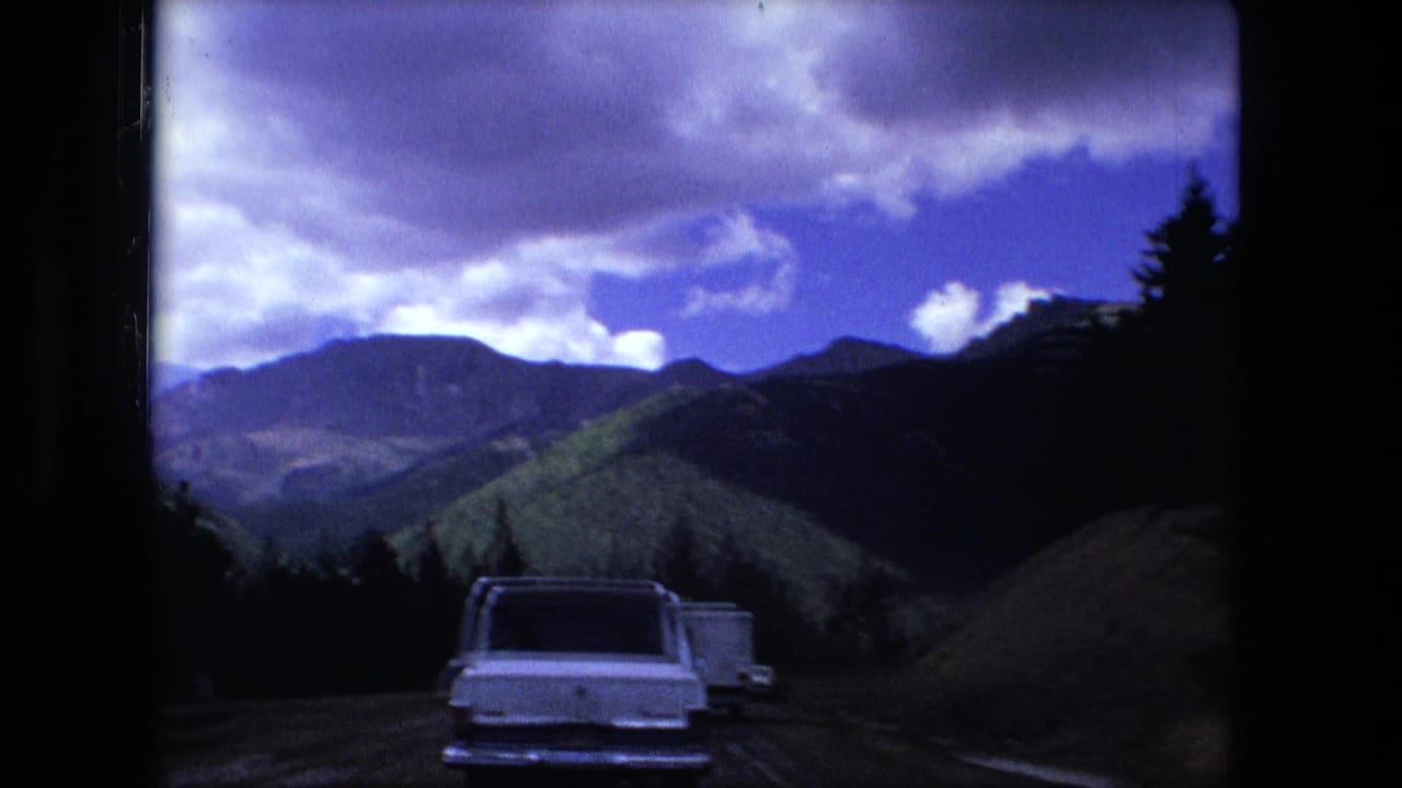 White car on a mountain road with forested slopes and clouds ahead.