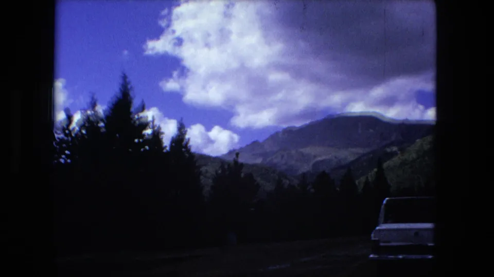 1976 8mm frame of a road through dark forest toward distant mountains, with a partial white car visible near the edge.