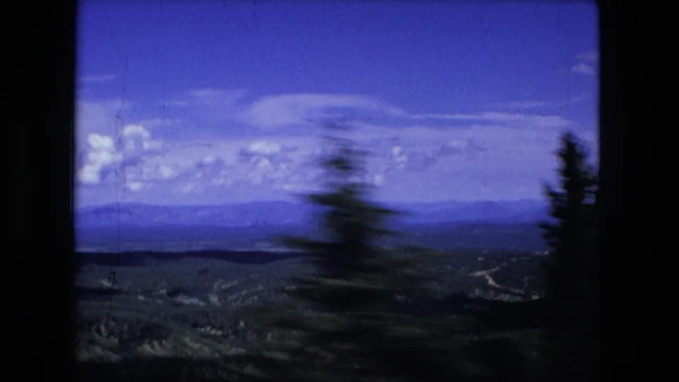 1976 8mm frame looking past a motion-blurred evergreen toward a wide valley, distant hills, and blue sky.