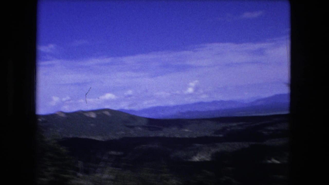 1976 8mm home-movie view of layered mountain ridges and a broad blue sky, framed by dark film borders.