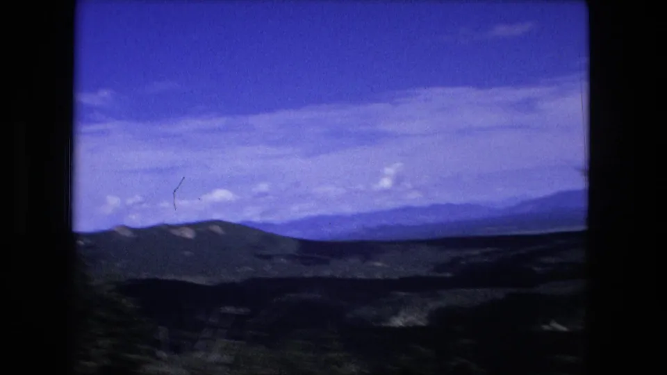 1976 8mm home-movie view of layered mountain ridges and a broad blue sky, framed by dark film borders.