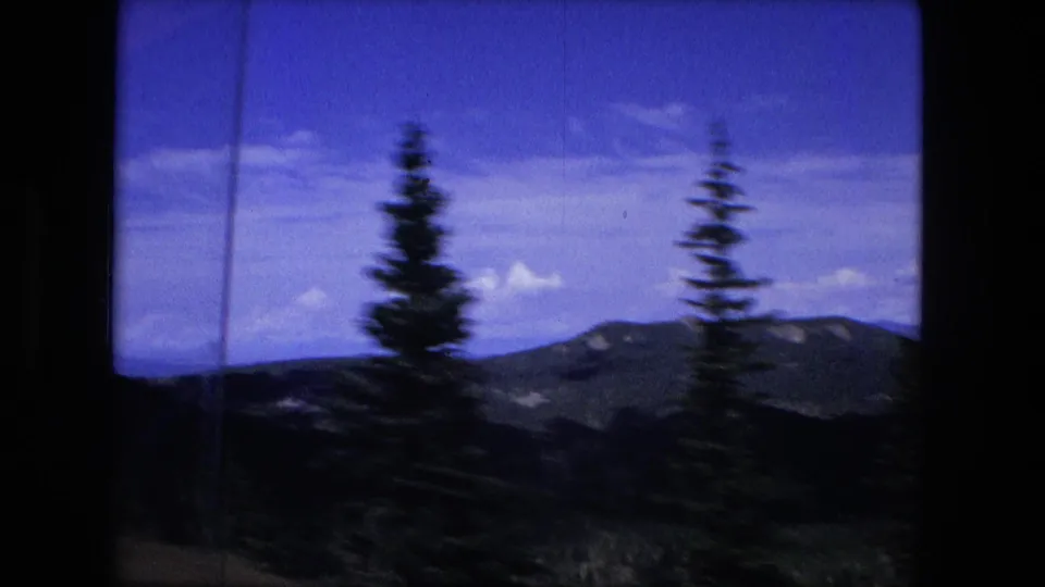 1976 8mm frame with pine trees in the foreground and low mountain ridges beyond, softened by travel motion.