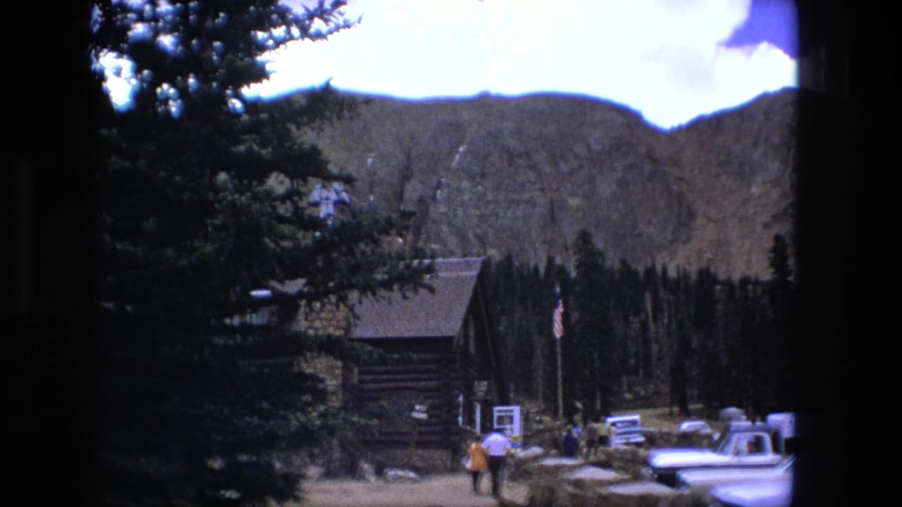 Log cabin and parked cars at the base of a mountain with visitors walking nearby.
