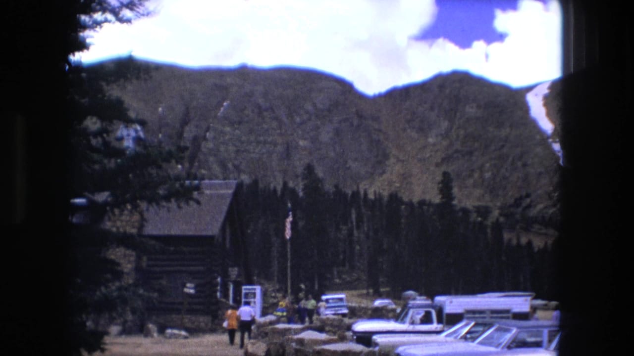 Rustic cabin, evergreen trees, visitors, and 1970s cars in a mountain parking area.
