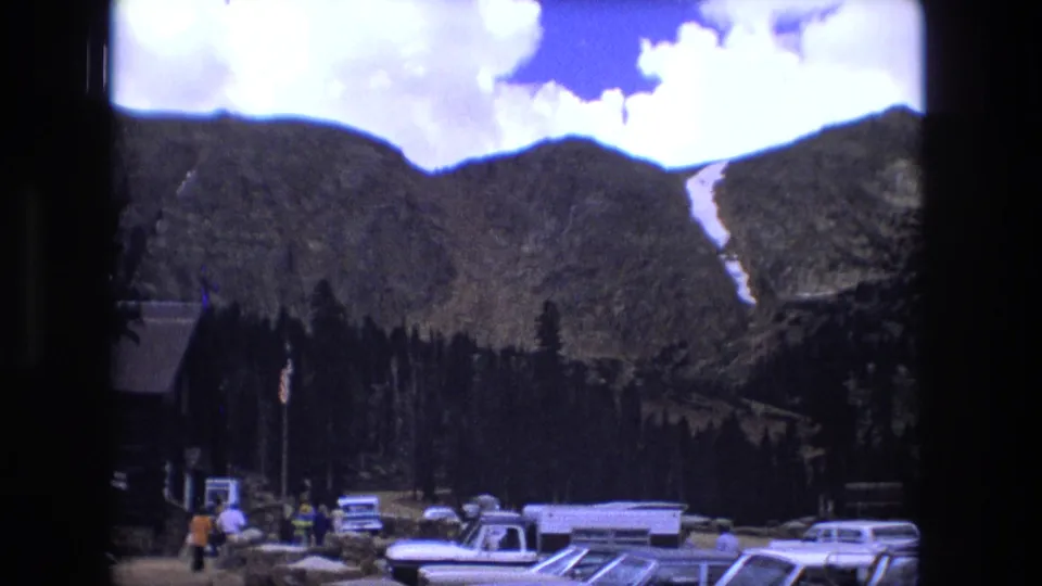 1976 home-movie frame looking across a mountain parking area with visitors, older cars, evergreens, and a snow patch on the slope.