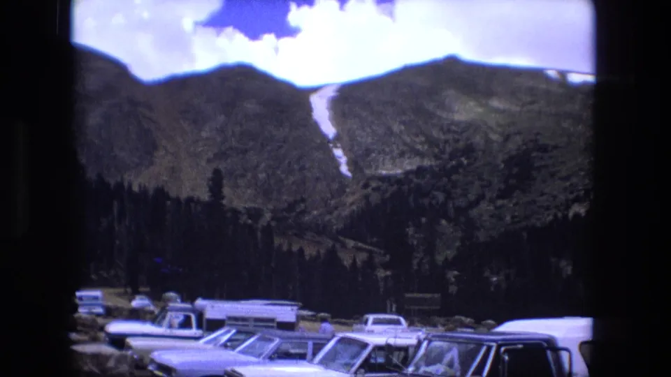 Final 1976 8mm frame showing period cars in the foreground, dark evergreen trees, and a rugged mountain slope with a narrow snow patch.
