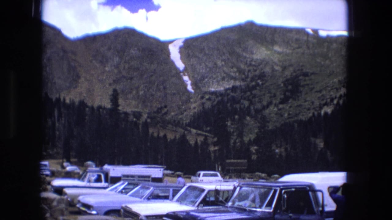 Mountain ridge above a parking lot with older cars and evergreen trees.