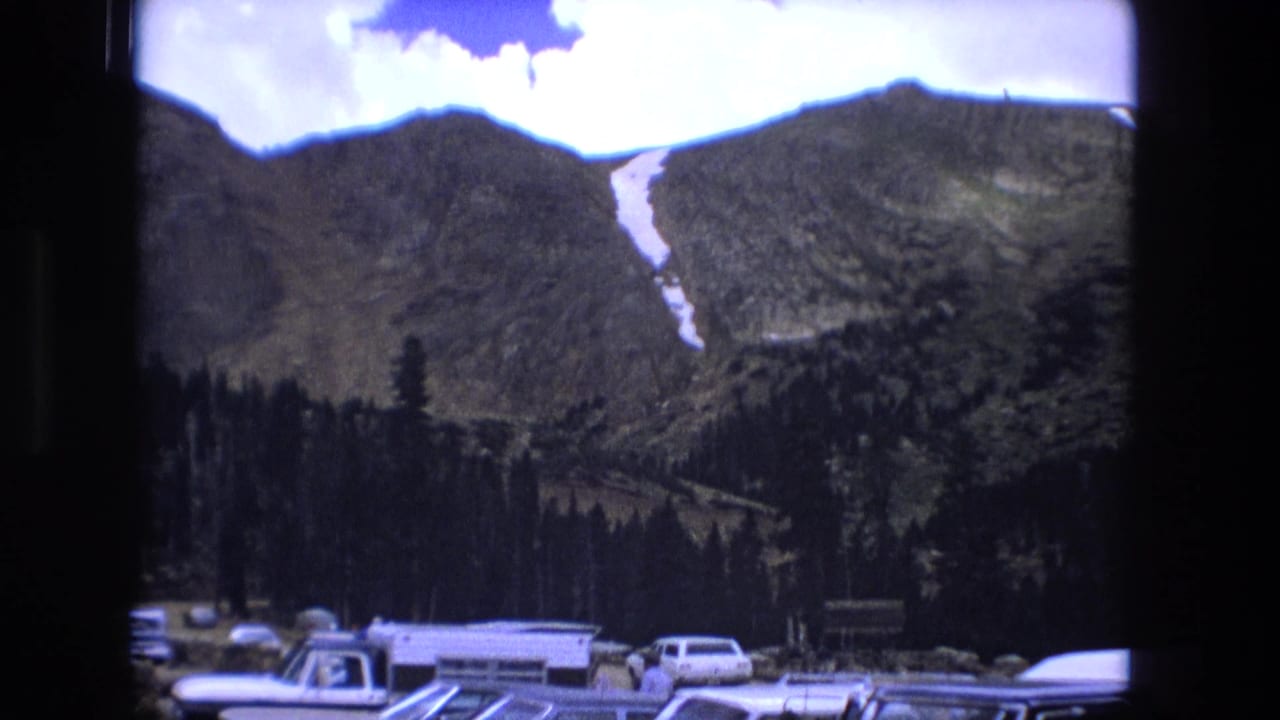 1976 8mm frame with parked vehicles in the foreground and a snow-marked mountain ridge behind dark evergreens.