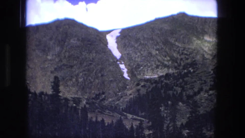 1976 8mm home-movie frame focused on a green mountain ridge with a lingering snow streak and dark evergreen foreground.