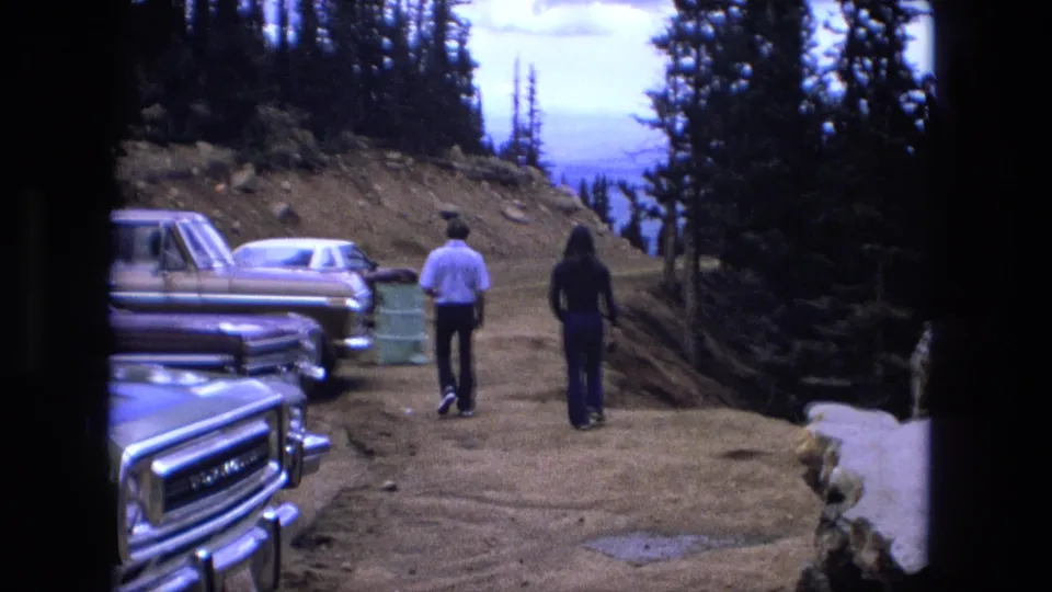 1976 8mm frame showing two people from behind walking away from a parking area toward a wooded overlook path.