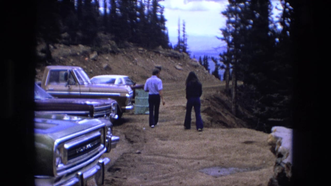 Two adults walk away from parked vehicles on a dirt mountain roadside.