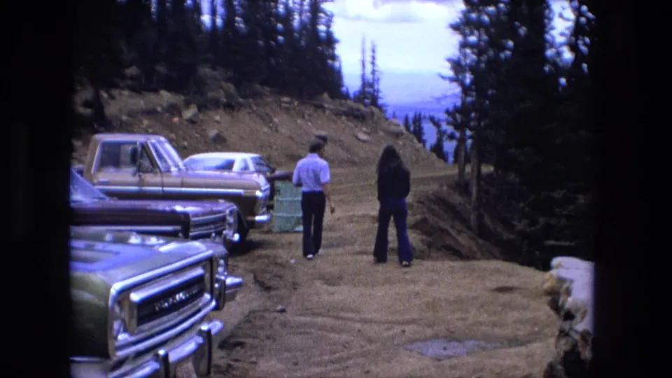 In 1970s 8mm footage, a man and woman walk from parked cars along a dirt mountain road toward evergreen trees and distant ridges.
