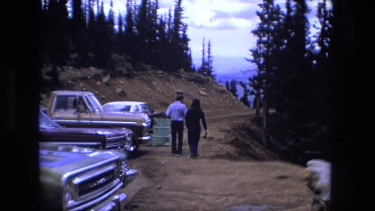 The couple continues along the dirt roadside, with older cars and a pickup parked near conifer trees and a distant mountain view.