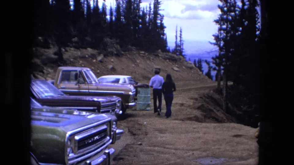 A handheld 8mm frame shows two adults moving away from classic cars and a pickup on a dirt mountain road under a bright sky.