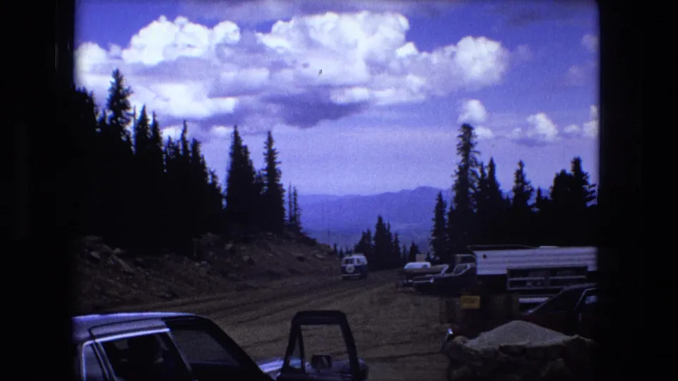 The final frame opens to a wider scenic view with parked vehicles, a trailer, evergreens, distant ridges, and a cloudy blue sky.