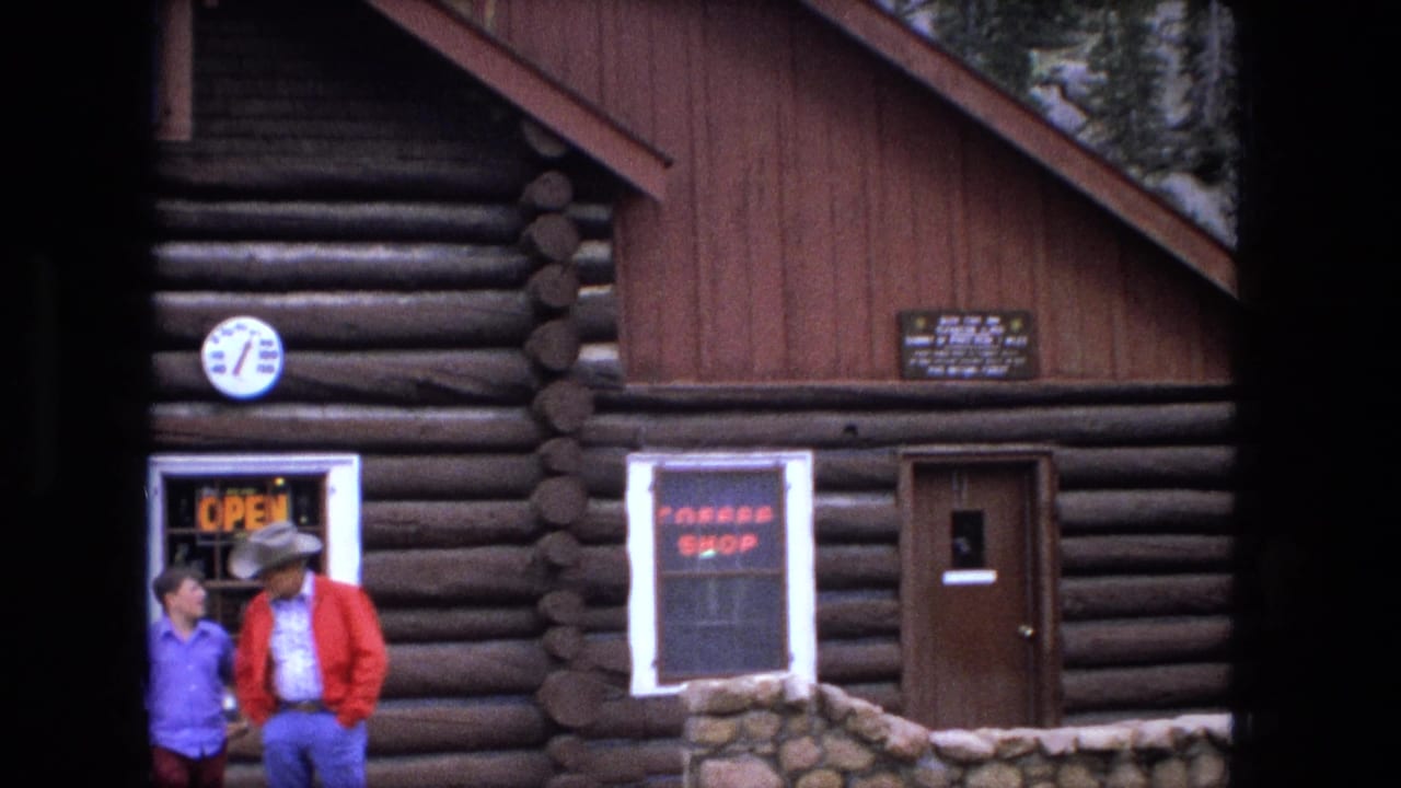 Two men stand outside a rustic log cabin with roadside signs.