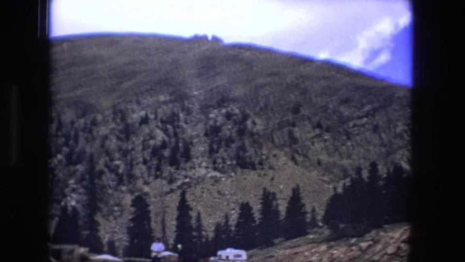 1976 8mm view of a rugged mountain slope with evergreen trees and a small white camper near the base.