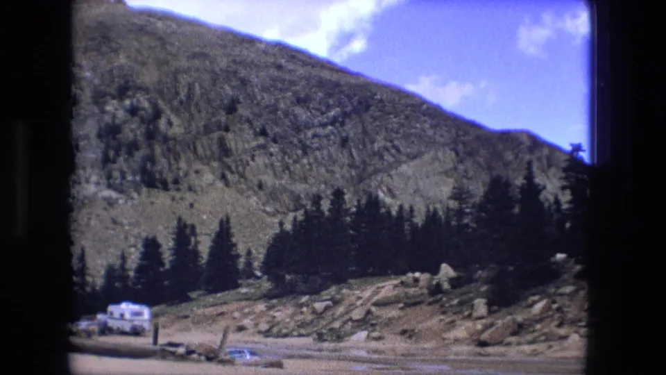 1976 8mm frame showing a mountain valley with dark evergreens, a white camper, parked cars, and rocky ground.