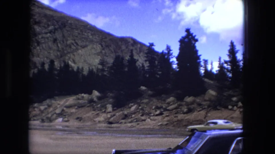 1976 8mm roadside view with cars at the lower edge, rocky ground, evergreen trees, and a mountain slope behind them.