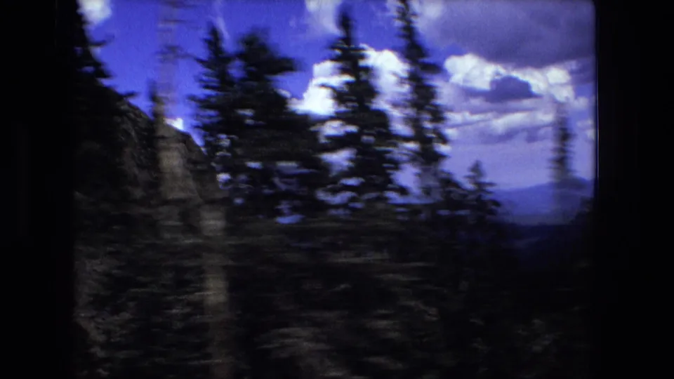 Handheld 8mm frame with dark evergreen trees and a rocky or wooded slope against a blue sky and far mountain horizon.