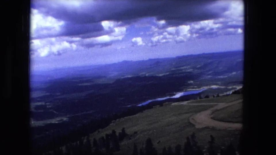 1970s 8mm view from a high slope showing a winding dirt road, scattered trees, distant hills, and a river or lake below.