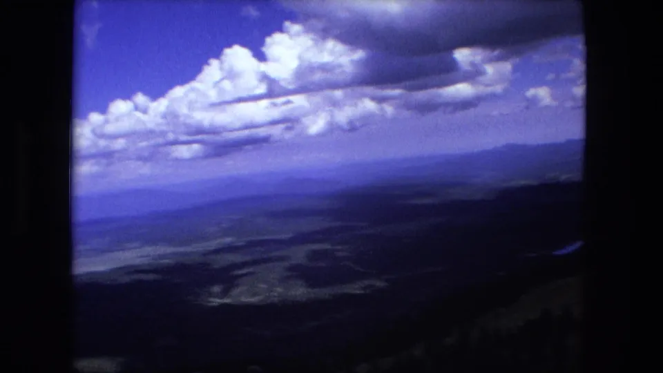 Soft 8mm frame looking over a broad valley with distant ridges, dark foreground slope, blue sky, and layered white clouds.