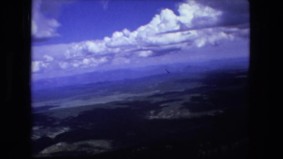 1976 8mm home-movie frame showing distant ridges, valleys, and cloud-filled blue sky from an aerial viewpoint.