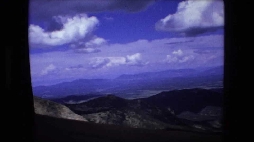 1976 8mm aerial frame with rolling ridges, a distant valley, and bright cumulus clouds above the horizon.
