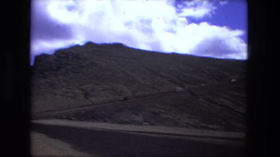 1976 8mm frame looking over a dark mountain slope with bright clouds and a narrow horizon line above it.