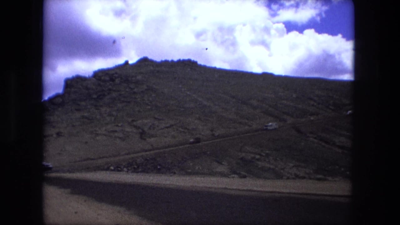 Mountain slope seen through a dark vehicle window under a bright clouded sky.