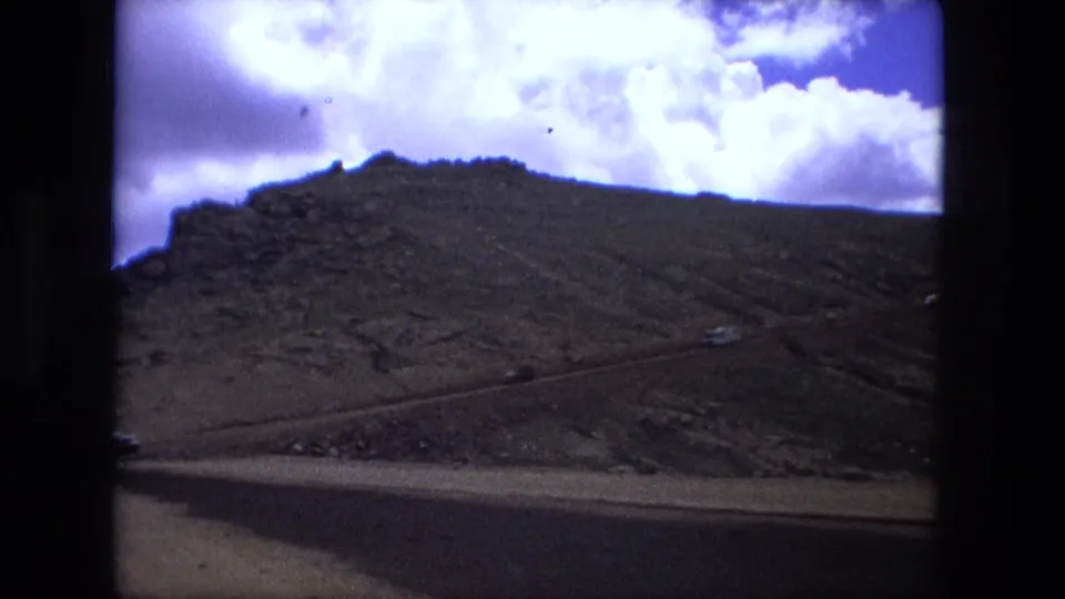 1970s 8mm-style view of rugged mountain terrain framed by a vehicle window, with blue sky and white clouds above.
