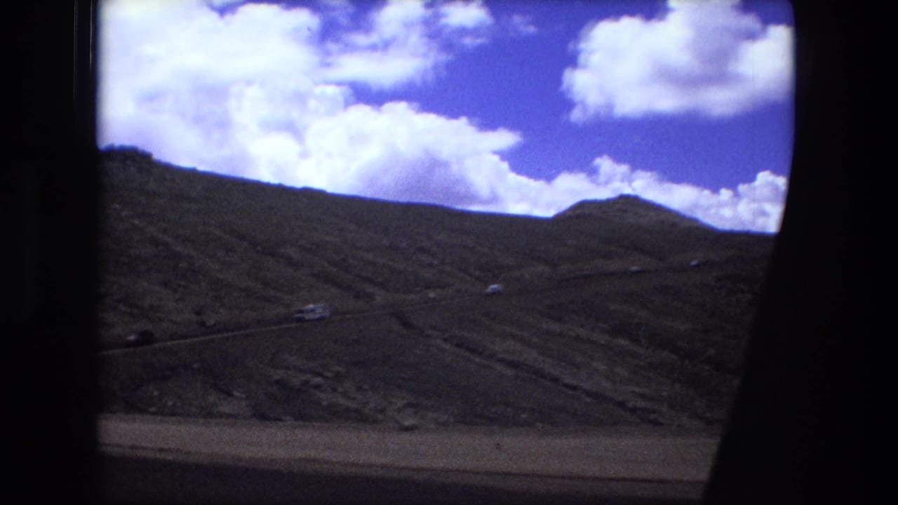 Mountain terrain and scattered clouds appear through a dark car-window frame in 1970s archival home-movie footage.