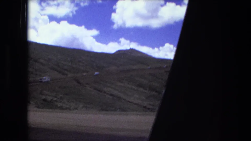 A sloped mountain landscape, road or track, and cloud-filled sky are framed by the vehicle interior in a home-movie travel shot.