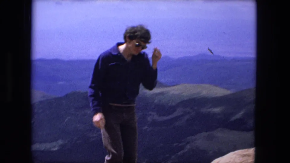 An adult man in sunglasses and dark clothing walks in a mountainous setting, with distant ridges and possible water or haze beyond.
