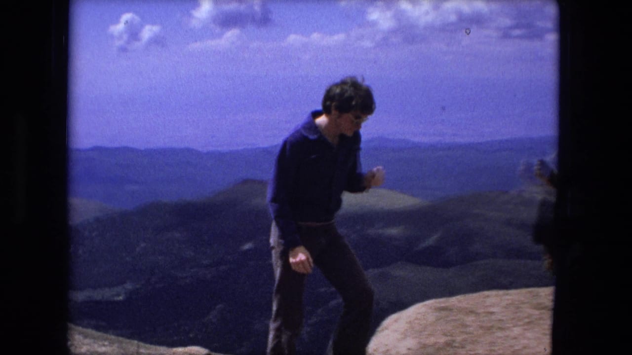 Young man in dark clothing walks on a rocky summit with distant hills behind him.