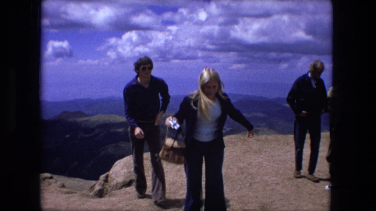 1976 8mm home-movie frame showing adults on a rocky mountain overlook, including a blonde woman in flared pants and a man in sunglasses.