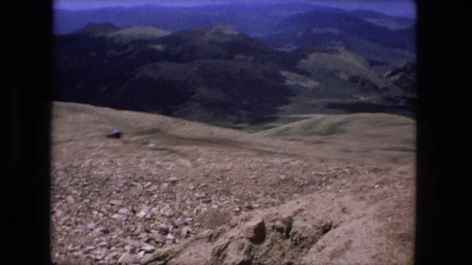 1976 8mm home-movie frame with a wide mountain and valley view from a rocky summit edge.