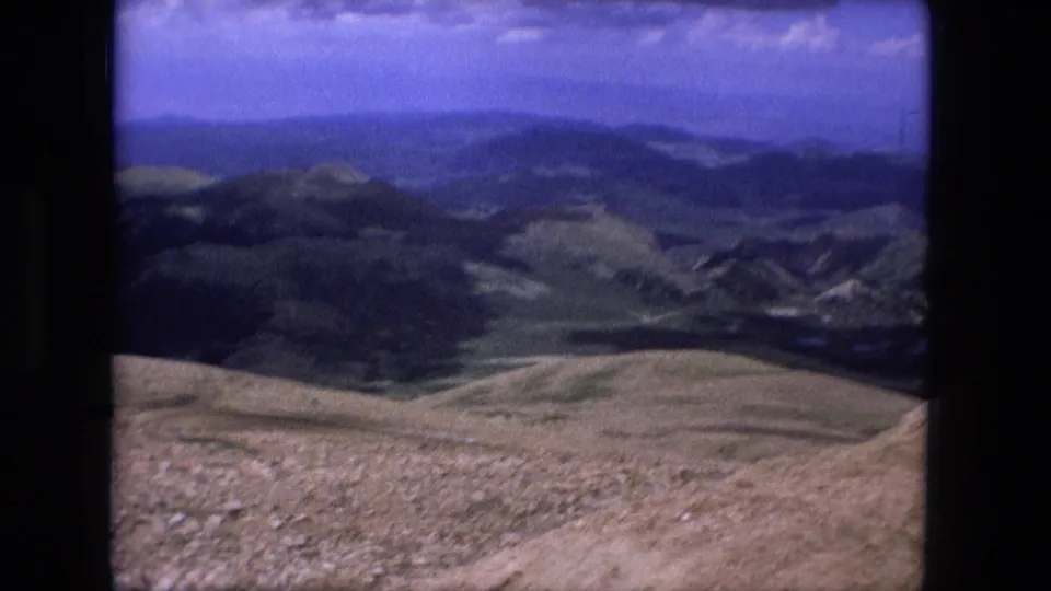 1976 8mm home-movie frame showing a panoramic mountain view with rocky ground, valleys, and layered hills under a blue sky.