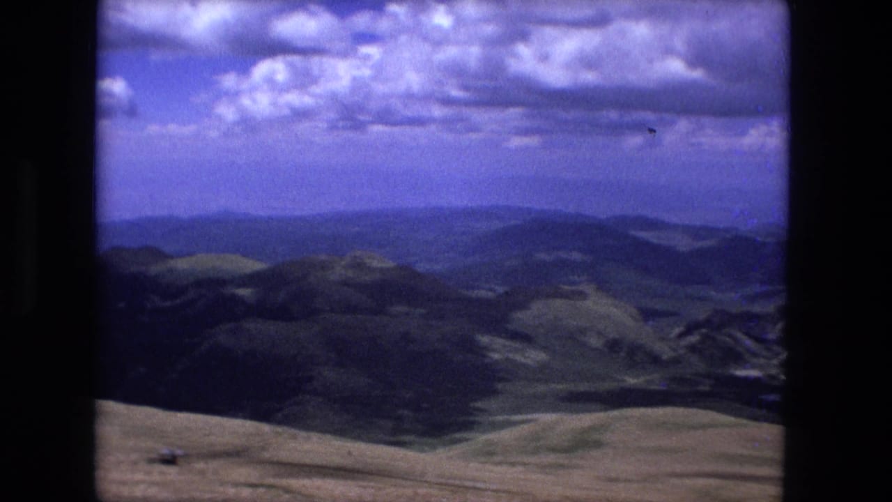 A 1970s 8mm landscape view shows layered mountain forms, pale foreground grassland, and distant blue haze beneath scattered clouds.