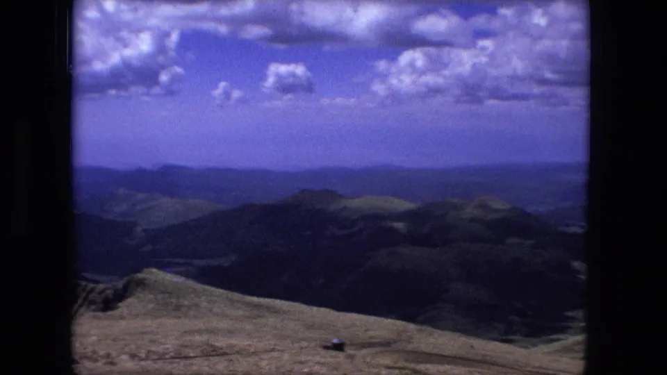 The frame holds a broad mountain view with dark ridges, pale open slope, distant horizon, and soft blue 8mm color cast.