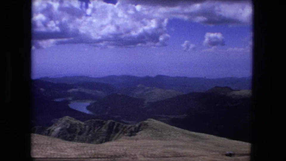 A 1976 8mm frame shows a high-country valley, distant ridges, and a small lake or winding river beneath a bright cloud pattern.