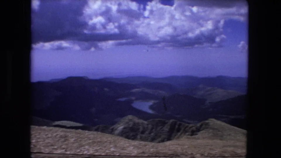 1976 8mm home-movie frame of layered mountain ridges, rocky foreground, and distant winding water beneath textured blue clouds.