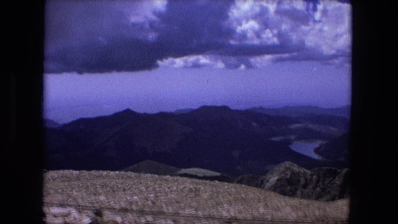 Cataloged 1976 8mm frame showing a high rocky overlook, layered ridges, distant water, and heavy clouds across a blue sky.