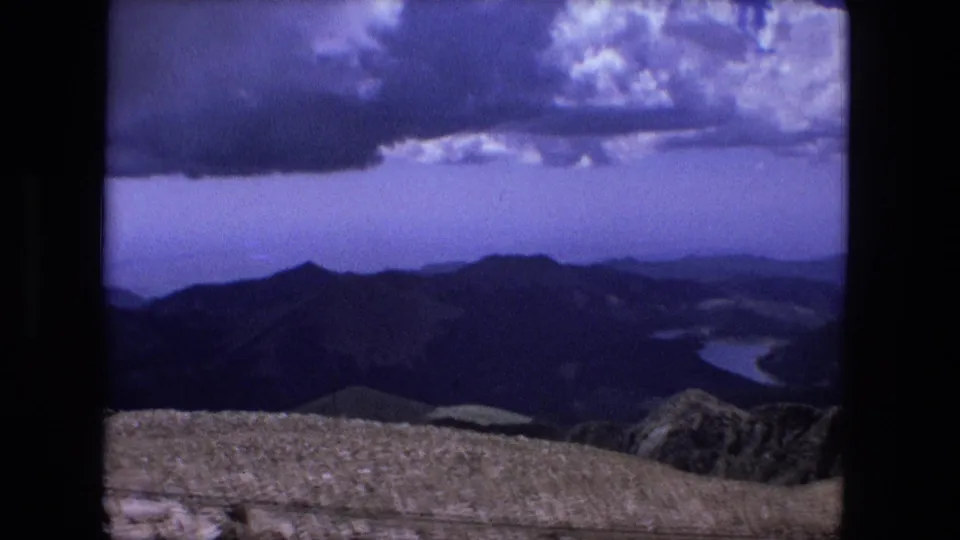 Cataloged 1976 8mm frame showing a high rocky overlook, layered ridges, distant water, and heavy clouds across a blue sky.