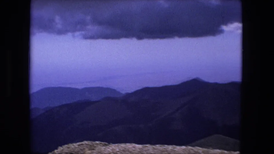 1976 8mm home-movie frame of hazy mountain layers beneath a dark cloud edge, with pale rocky ground at the bottom.